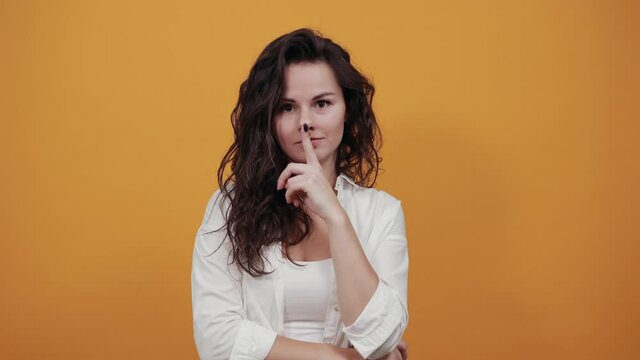 Index finger on lips, silence gesture, shhh quiet, asks for voicelessness forefinger at the mouth. Young attractive woman, dressed white blouse, with brown eyes, curly hair, yellow background