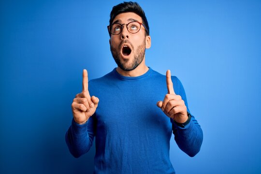Young handsome man with beard wearing casual sweater and glasses over blue background amazed and surprised looking up and pointing with fingers and raised arms.