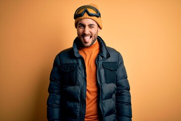 Young handsome skier man with beard wearing snow sportswear and ski goggles sticking tongue out happy with funny expression. Emotion concept.