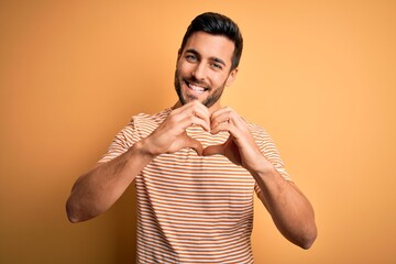 Young handsome man with beard wearing casual striped t-shirt over yellow background smiling in love doing heart symbol shape with hands. Romantic concept.