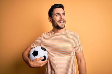 Handsome player man with beard playing soccer holding footballl ball over yellow background looking away to side with smile on face, natural expression. Laughing confident.