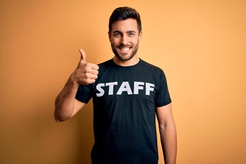 Young handsome worker man with beard wearing staff uniform t-shirt over yellow background happy...