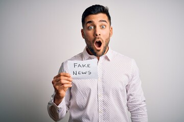 Young business man holding fake news paper over isolated background scared in shock with a surprise...