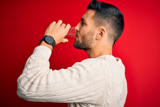 Young handsome man drinking glass of healthy water to refreshment standing over isolated red background
