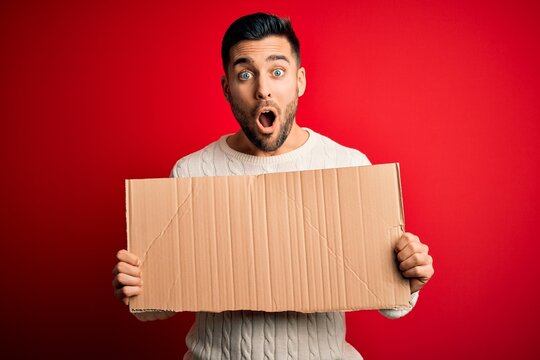 Young handsome man holding blank cardboard banner over isolated red background scared in shock with a surprise face, afraid and excited with fear expression
