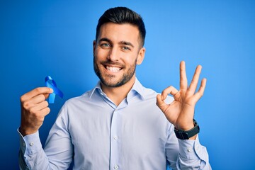 Young handsome man holding blue ribbon as prostate campaing support over blue background doing ok...