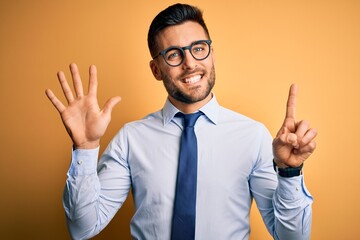 Young handsome businessman wearing tie and glasses standing over yellow background showing and pointing up with fingers number six while smiling confident and happy.