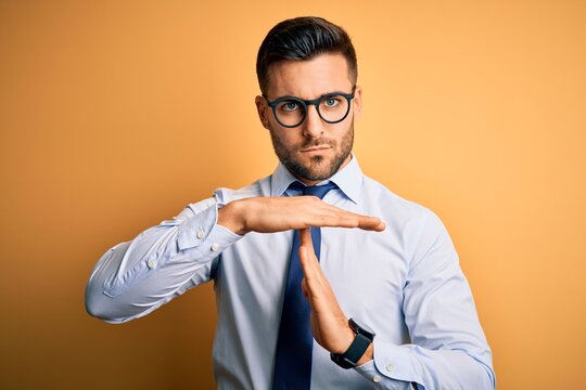 Young handsome businessman wearing tie and glasses standing over yellow background Doing time out gesture with hands, frustrated and serious face