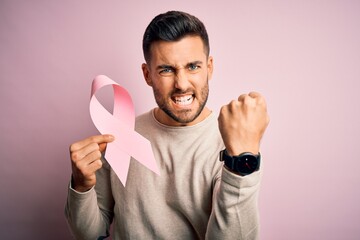 Young handsome man holding pink cancer ribbon symbol of support over isolated background annoyed...