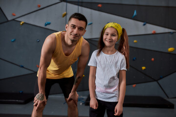 Young male instructor and active little girl looking at camera, standing against artificial training climbing wall. Concept of sport life and rock climbing