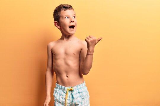 Cute blond kid wearing swimwear pointing thumb up to the side smiling happy with open mouth