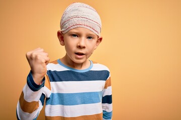 Young little caucasian kid injured wearing medical bandage on head over yellow background angry and mad raising fist frustrated and furious while shouting with anger. Rage and aggressive concept.