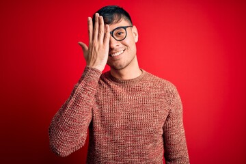 Young handsome hispanic man wearing nerd glasses over red background covering one eye with hand, confident smile on face and surprise emotion.