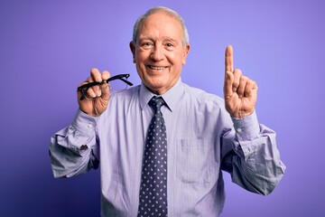Senior grey haired man holding correction glasses over purple background surprised with an idea or question pointing finger with happy face, number one