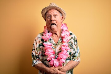 Grey haired senior man wearing summer hat and hawaiian lei over yellow background sticking tongue...