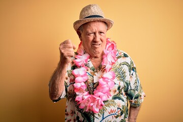 Grey haired senior man wearing summer hat and hawaiian lei over yellow background angry and mad...