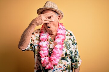 Grey haired senior man wearing summer hat and hawaiian lei over yellow background peeking in shock...