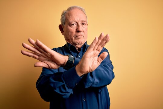 Grey haired senior man wearing casual blue shirt standing over yellow background Rejection expression crossing arms doing negative sign, angry face