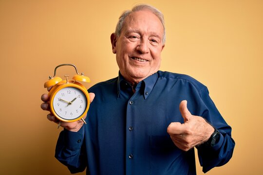 Senior grey haired man holding vintage alarm clock over yellow background happy with big smile doing ok sign, thumb up with fingers, excellent sign