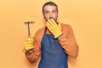 Young handsome man wearing gardener apron and gloves holding rake covering mouth with hand, shocked...