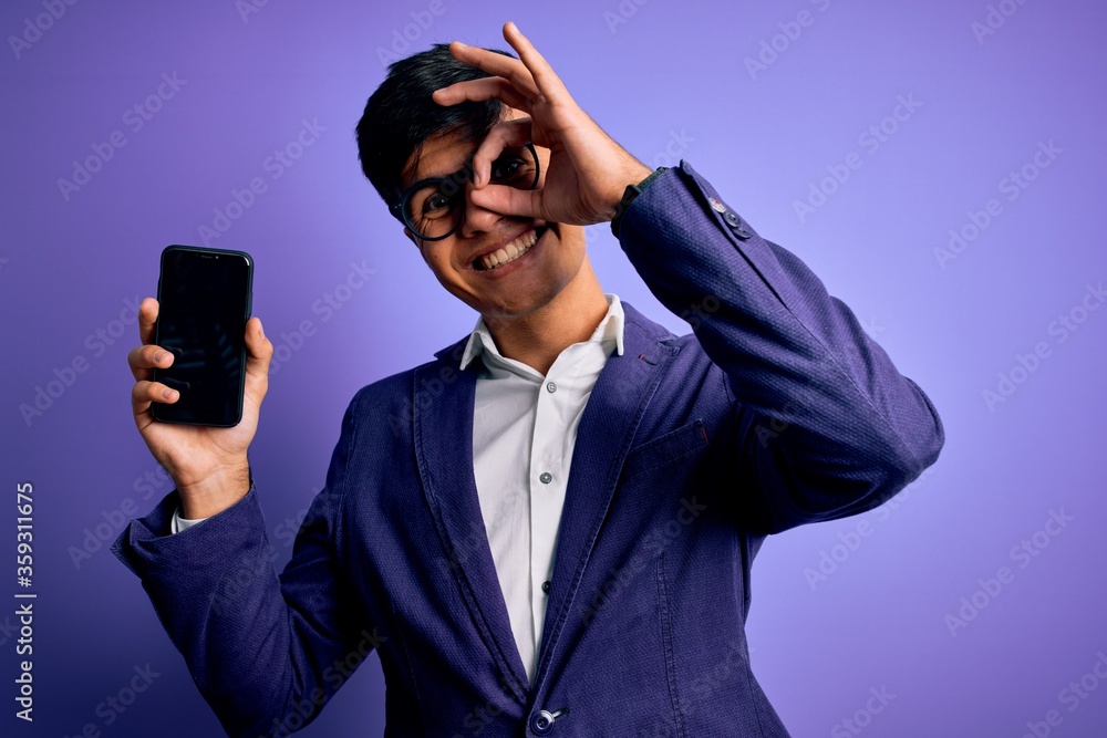 Poster Young handsome business man wearing jacket and glasses holding smartphone showing screen with happy face smiling doing ok sign with hand on eye looking through fingers