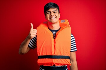 Young handsome man wearing orange safety life jacket over isolated red background doing happy...