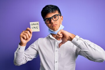 Young handsome man wearing medical mask holding reminder with corona virus message with angry face, negative sign showing dislike with thumbs down, rejection concept