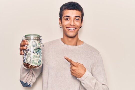 Young african amercian man holding jar with savings smiling happy pointing with hand and finger