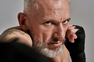 Close up of middle aged athletic man, kickboxer looking agressive while boxing, training in studio over grey background. Muay Thai, Boxing or Kickboxing concept