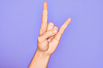 Hand of caucasian young man showing fingers over isolated purple background gesturing rock and roll symbol, showing obscene horns gesture