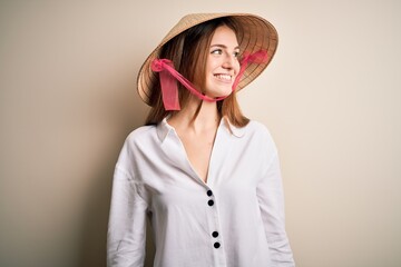 Young beautiful redhead woman wearing asian traditional conical hat over white background looking away to side with smile on face, natural expression. Laughing confident.