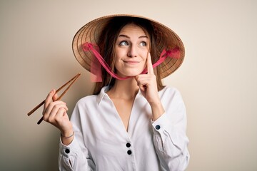 Young beautiful redhead woman wearing asian traditional hat holding wooden chopsticks serious face...