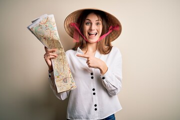 Young beautiful redhead woman wearing asian traditional hat holding city map very happy pointing with hand and finger