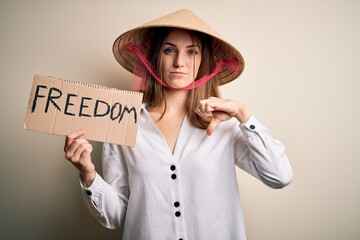 Young redhead woman wearing asian traditional hat holding banner with freedom message with angry...