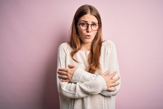 Young beautiful redhead woman wearing casual sweater and glasses over pink background shaking and freezing for winter cold with sad and shock expression on face