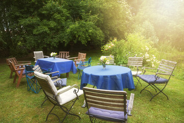 Set up for a private garden party with tables, blue tablecloths and many different chairs in a...