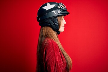 Young beautiful redhead motocyclist woman wearing moto helmet over red background looking to side, relax profile pose with natural face with confident smile.