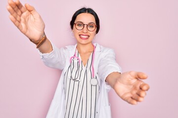 Beautiful doctor woman with blue eyes wearing coat and stethoscope over pink background looking at...