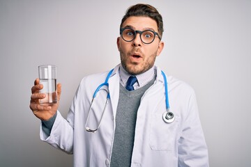 Young doctor man wearing medical coat holding a glass of fresh water over isolated background scared in shock with a surprise face, afraid and excited with fear expression