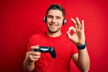 Young gamer man with blue eyes playing video games using gamepad joystick doing ok sign with fingers, excellent symbol