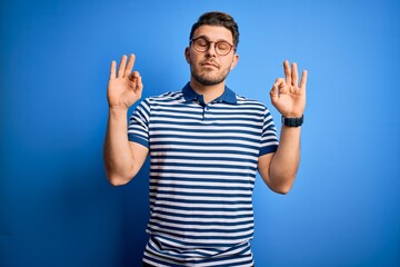 Young man with blue eyes wearing glasses and casual striped t-shirt over blue background relax and smiling with eyes closed doing meditation gesture with fingers. Yoga concept.