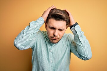 Young business man with blue eyes wearing elegant green shirt over yellow background suffering from headache desperate and stressed because pain and migraine. Hands on head.