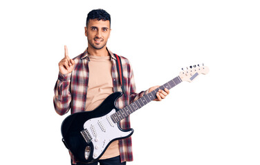 Young hispanic man playing electric guitar surprised with an idea or question pointing finger with happy face, number one