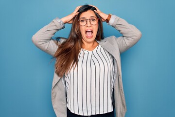 Young hispanic business woman wearing glasses standing over blue isolated background Crazy and scared with hands on head, afraid and surprised of shock with open mouth