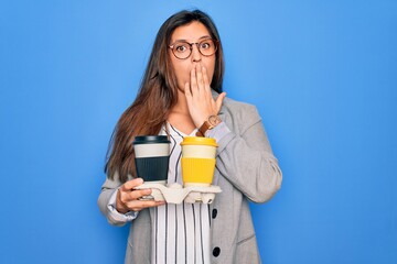 Young hispanic intern business woman holding coffee over blue background cover mouth with hand shocked with shame for mistake, expression of fear, scared in silence, secret concept
