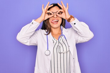 Professional doctor woman wearing stethoscope and medical coat over purple background doing ok gesture like binoculars sticking tongue out, eyes looking through fingers. Crazy expression.