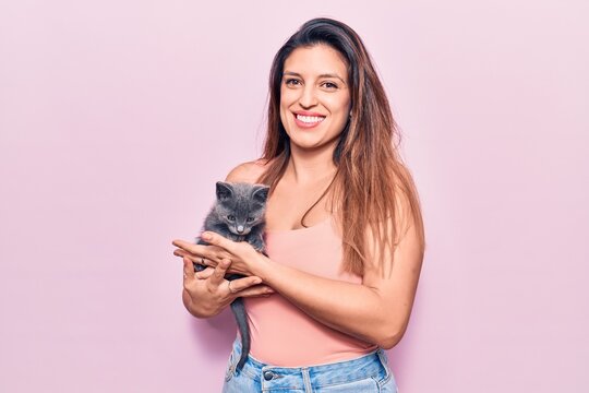 Young beautiful latin woman smilling happy. Standing with smile on face holding adorable cat over isolated pink background