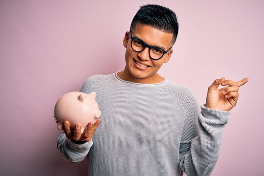 Young handsome latin man holding piggy bank over isolated pink background very happy pointing with hand and finger to the side