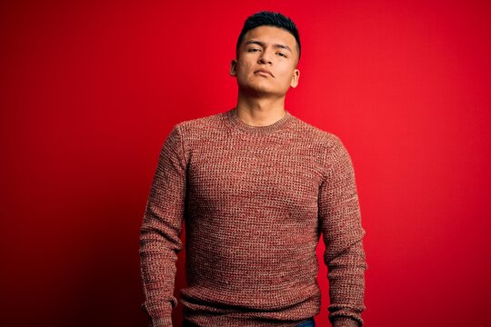 Young handsome latin man wearing casual sweater standing over red background Relaxed with serious expression on face. Simple and natural looking at the camera.