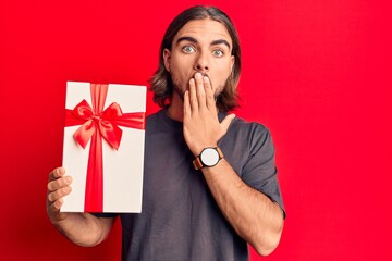 Young handsome man holding gift covering mouth with hand, shocked and afraid for mistake. surprised...